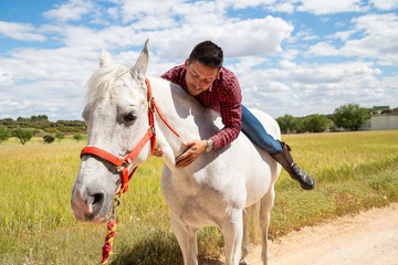 Excited young guy in casual outfit smiling and embracing neck of white horse during ride in field on cloudy day
