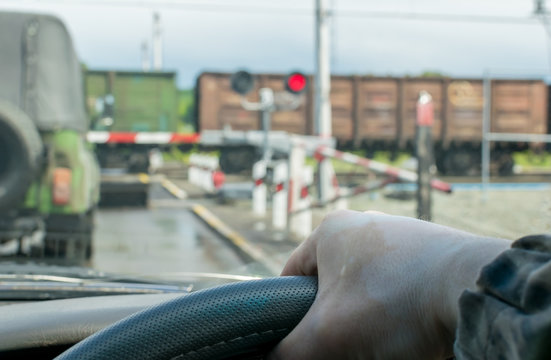 View Of The Driver Hand On The Steering Wheel Of The Car, Which Stopped In Front Of A Closed Railway Crossing At A Red Traffic Light. The Driver Waits For The Freight Train To Pass