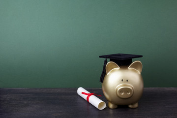 Gfold piggy bank with a grad cap and diploma in front of green chalkboard. Education scholarship