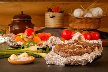 Meat steak baked in foil with fresh herbs and vegetables on a wooden table against a background of rustic utensils