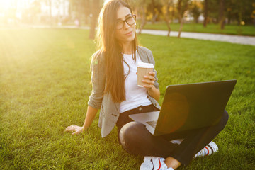 Image of beautiful stylish woman sitting on green grass with laptop and coffee in the hand. She is talking on the phone through wireless headphones. Sunset light. Lifestyle concept