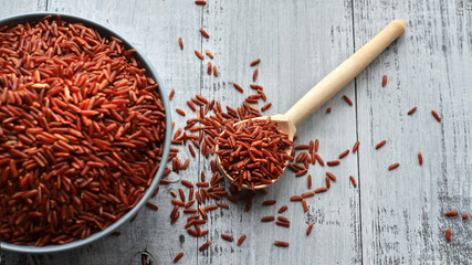 Bowl and full spoon with uncooked red rice on wooden background, top view