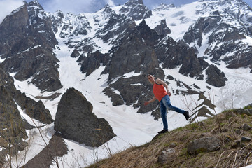 Athletic young man in the orange shirt on top of snow and the rocky mountains on a warm Sunny spring day.