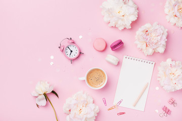 Coffee, macaroons, alarm clock, office supply, peony flowers and notebook on pink pastel table top view. Flat lay. Beautiful morning breakfast. Fashion woman working desk.