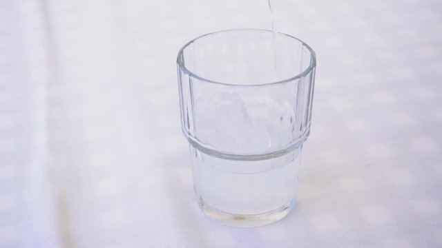 Serving ouzo with ice cubes on a small glass. Pouring strong alcoholic drink ouzo on the rocks, served on a glass on a Greek tavern table with white linen tablecloth.