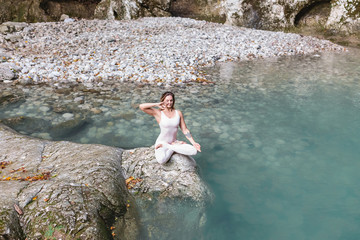 Filling with energy and inspiration from nature. Beautiful young female yoga coach meditates and relaxes while sitting in the lotus position on a stone near a clean mountain lake