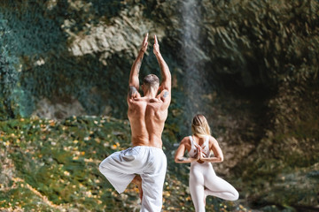Couple - beautiful man and female advanced yoga instructors are engaged in nature standing barefoot on rocky beach next to mountain river against picturesque green hills. Concept of unity with nature