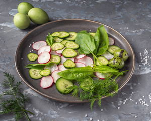 A large plate of sliced radish and cucumber, fresh spinach seasoned with salt and pepper. In the background are three boiled eggs, painted in green. Sprigs of fresh dill.