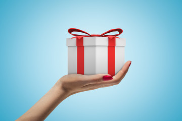 Side closeup view of woman's hand holding white gift box tied with red ribbon on light blue background.