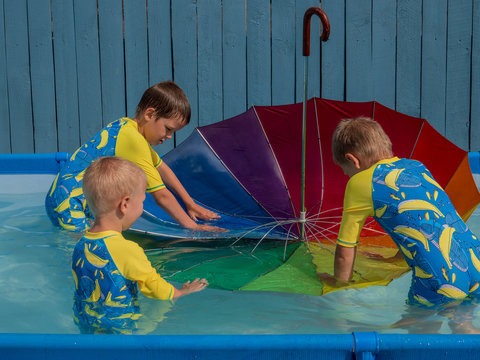 happy kids playing in carcas pool in boys' swimsuits with rainbow umbrella. brothers are happy together in warm water on sunny summer day. concept of educational games for preschoolers