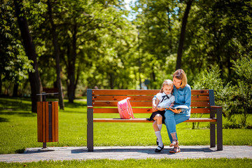 Fototapeta premium Mother and little daughter in outdoors public park sitting on the bench and read book, study lessons. Parenthood and child concept.