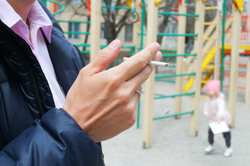 man smokes near children playground, hand with a cigarette and smoke closeup, bad habits concept