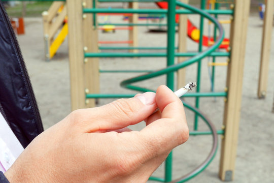 man smokes near children playground, hand with a cigarette and smoke closeup, bad habits concept
