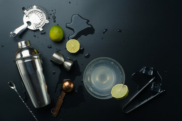 Margarita cocktail and bar equipments, stainless steel cocktail shaker and jigger, copper measuring spoon, bar spoon with strainer, the lemons and ice tongs with ice cubes on the table