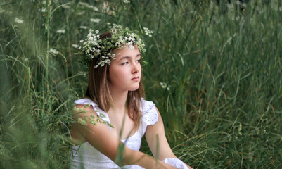 Beautiful romantic preteen girl with green and white fresh flowers head wreath sitting in green grass at wild meadow outdoors. Proximity to nature. Natural beauty