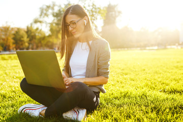 Image of beautiful stylish woman sitting on green grass with laptop  She is talking on the phone through wireless headphones. Sunset light. Lifestyle concept.