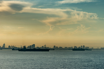 Makassar, Sulawesi, Indonesia - February 28, 2019: Early morning yellowish light over city skyline fronted by a number of dark large ships on Flores Sea, under cloudscape.