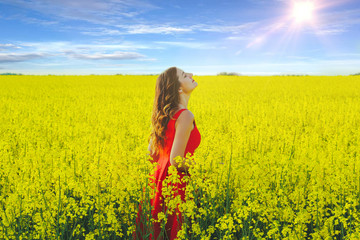 Fototapeta premium Young beautiful girl in a red dress close up in the middle of yellow field with radish flowers and sunlight..