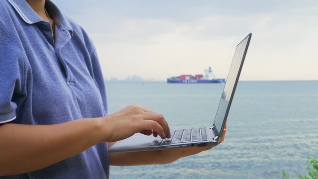 Close up shot of young woman foreman using laptop for check product at commercial port, shipping vessels that are carrying goods in blurred background.