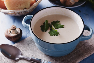 Creamy mushroom soup with leaf of cilantro in a blue ceramic bowl next to row of fresh mushrooms and spoon on a blue wooden background, closeup