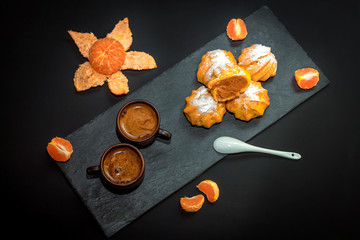 Fresh hot coffee in ceramic cups, ripe tangerine and sweet dessert – profiterole or eclair cakes...