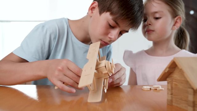 Brother and sister assemble a dinosaur 3d puzzle of wooden parts. The girl gives her brother details and watches with interest the process. Studio video shot with dolly pan of child hobby in 4K