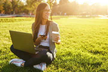 Image of beautiful stylish woman sitting on green grass with laptop and phone in the hands. She is talking on the phone through wireless headphones. Sunset light. Lifestyle concept