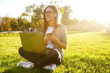 Naklejka na ściany i meble Image of beautiful stylish woman sitting on green grass with laptop and phone in the hands. She is talking on the phone through wireless headphones. Sunset light. Lifestyle concept