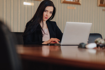 Young attractive emotional girl in business-style clothes sitting at a desk on a laptop and phone in the office or auditorium