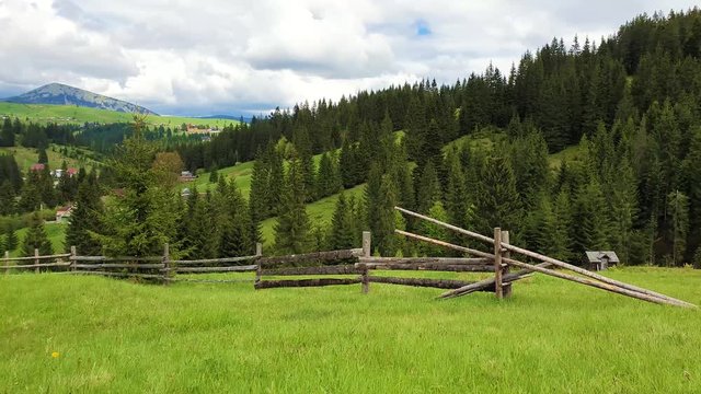 Picturesque spring mountains scene with wooden split rail fence across a green and lush pasture with a old house on the valley surrounded by pine woods.