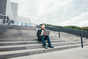 Attractive adult successful bald bearded man in black jacket using laptop in stairs at city