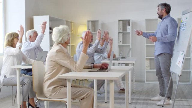 Side view of male teacher with beard standing before whiteboard and teaching computer skills to group of elderly people sitting at desks and raising hands to answer question