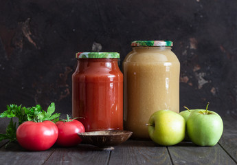 Jars with homemade tomato and apple juice, fresh tomatoes, parsley, salt and apples on a dark wooden background.