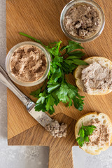 Liver pate in a glass jar with fresh bread and parsley on a wooden cutting board. Light grey stone background.