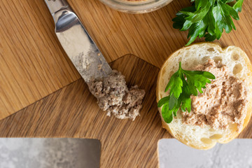 Liver pate in a glass jar with fresh bread and parsley on a wooden cutting board. Light grey stone...