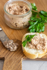Liver pate in a glass jar with fresh bread and parsley on a wooden cutting board. Light grey stone background.