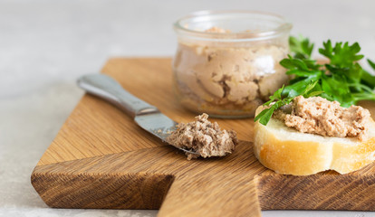 Liver pate in a glass jar with fresh bread and parsley on a wooden cutting board. Light grey stone background.
