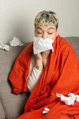 Unhealthy girl sitting on the couch, wrapped in a warm orange blanket, holding a napkin, sneezing...