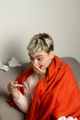 Unhealthy girl sitting on the couch, wrapped in a warm orange blanket, holding a napkin, sneezing and blowing her nose. Girl looks with chronic allergic rhinitis or cold difficult to breathe concept.