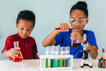 Two African American mixed kids testing chemistry lab experiment and holding glass tube flask along with microscope on table and smile in science classroom - fun learning education concept