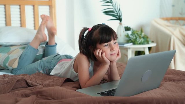 Little girl lying on bed and watching cartoon on laptop in her room, slow motion