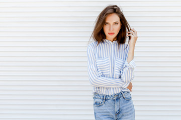 Young stylish beautiful brunette woman wearing blue striped shirt and denim jeans posing outdoors on a background of white wall. Trendy casual spring summer outfit. Street fashion. Space for text.