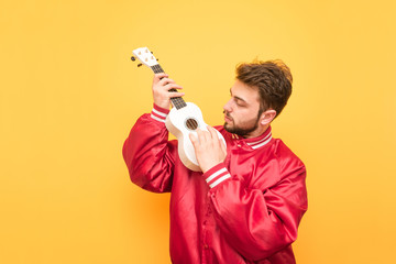 Man in a red jacket plays a Hawaiian guitar on the background of a yellow wall. Student with a beard is isolated on a yellow background with a ukulele in his hand. Musical concept