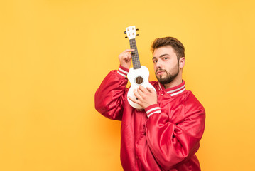 Fototapeta na wymiar Portrait of an adult man standing with an ukulele in his hands on a yellow background and posing on a camera wearing a red jacket. Bearded man plays music on a Hawaiian guitar. Isolated