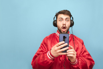 Surprised man with a beard in the headphones, holds a smartphone in his hands and looks at the camera with a shocking face. Emotional man listens to music and uses a smartphone on a blue background