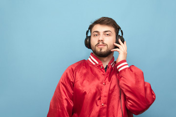 Portrait of a bearded man in his headphones and a red jacket on a blue background, looking in camera. Adult man listens to music in headphones. Isolated.