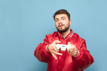 Funny man with a beard standing on a blue background with a gamepad in his hand and playing a video game, the beauty of a red jacket. Fun person playing video games on the console. isolated.