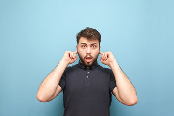 Amotional man with a surprised face closes his ears with his fingers and looks at the camera,...