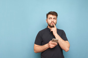 Thoughtful man with a beard standing on a blue background with a smartphone in his hand, looking...