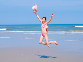 Obraz na płótnie Canvas Young happy teen girl having fun on tropical beach and jumping in pink swimsuit and striped hat into the air on the sea coast at the day time. Summer travel and vacation concept